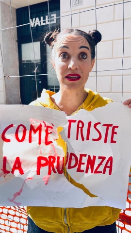 come è triste la prudenza, recita un cartello tenuto da un'attrice 
